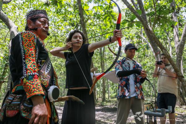 Liana shooting a bow and arrow, observed by the tour guide.